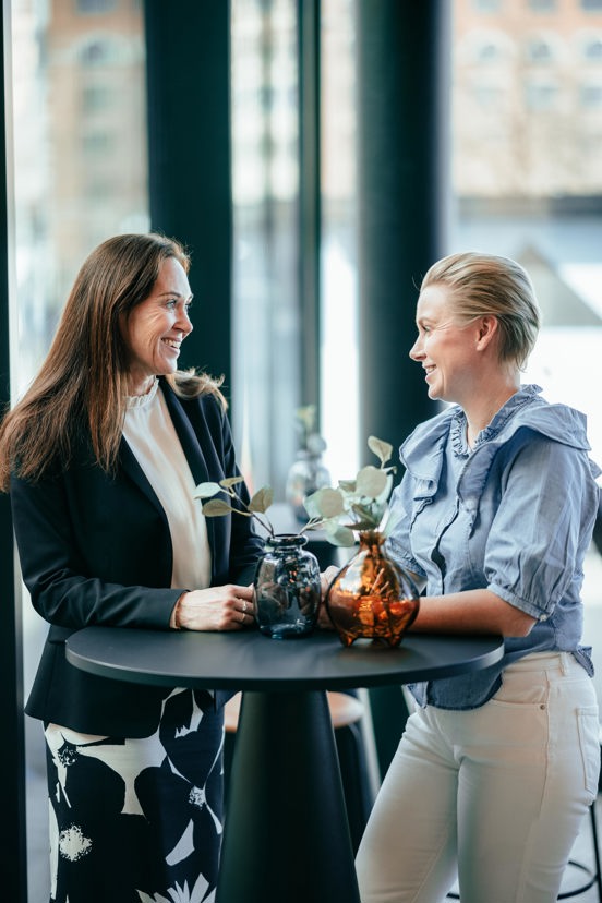 To damer står rundt et bord og snakker