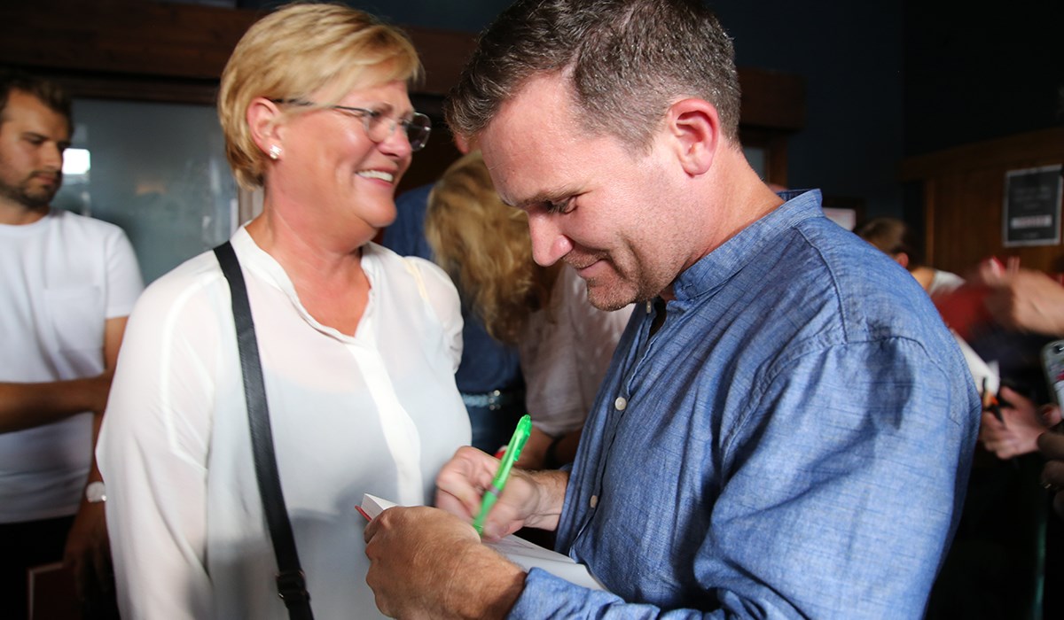 Bilde: Mann signerer bok stående med folk i bakgrunnen.