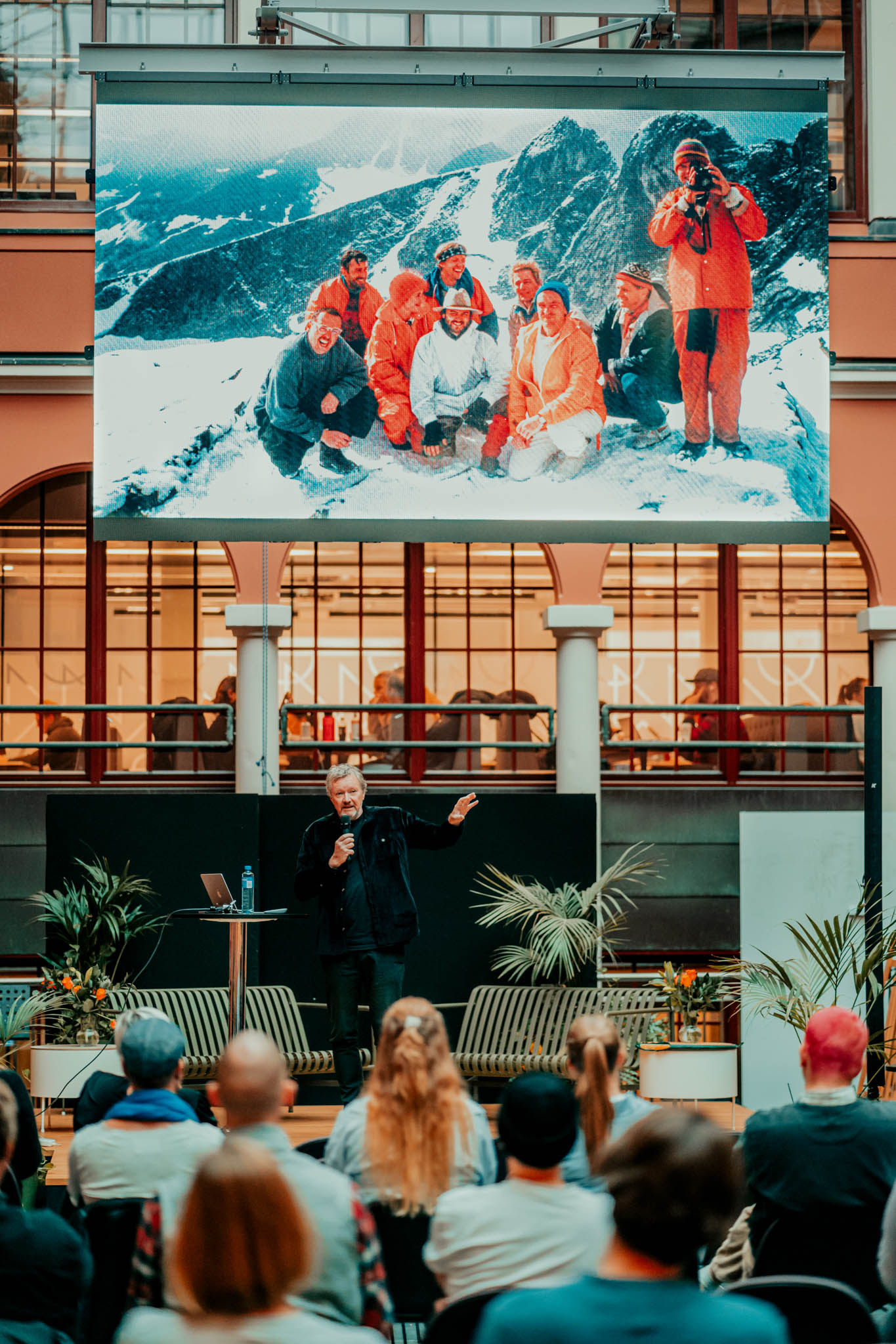 Kjetil Trædal Thorsen står på scenen foran forsamlingen mens han peker på en stort skjerm over han som viser en gjeng i orange klær på ekspedisjon på et snødekt fjell. Foto.