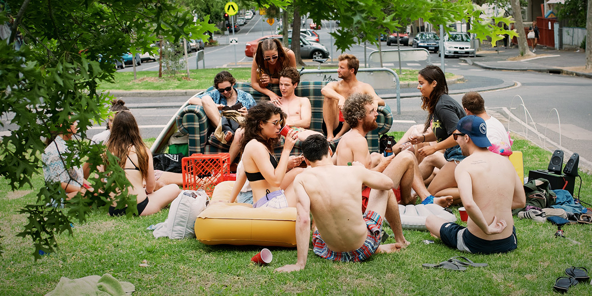 En gruppe studenter sitter på en gressplen. Det er en sofa plassert på gresset som også noen studenter sitter i. Studentene er kledd i sommerklær og noen av guttene har bar overkropp. 