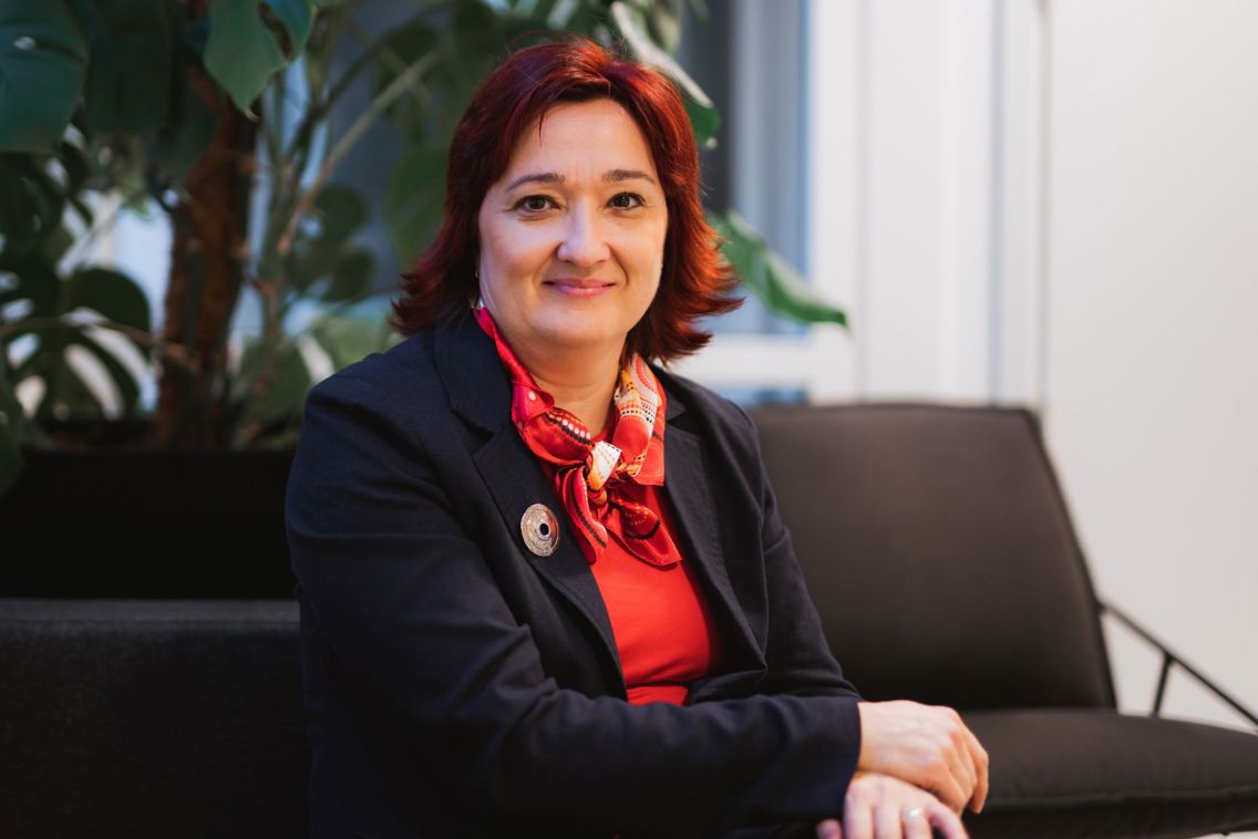 Miroslava sitter i mørk sofa og ser inn i kamera, med plante i bakgrunnen