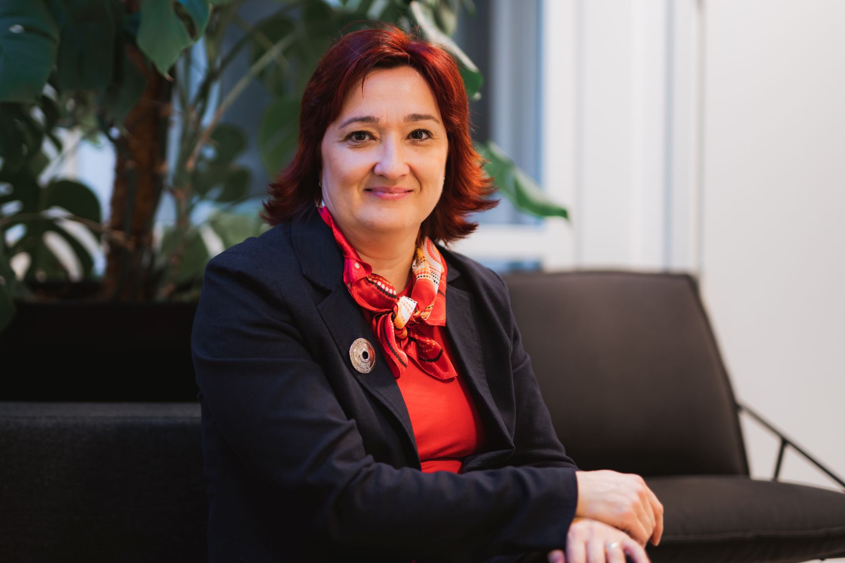 Miroslava sitter i mørk sofa og ser inn i kamera, med plante i bakgrunnen