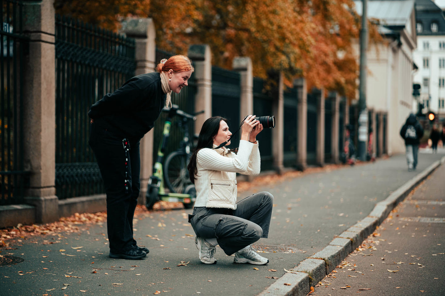 To Digital innholdsproduksjon-studenter står på et fortau og fotograferer noe utenfor bilderammen.