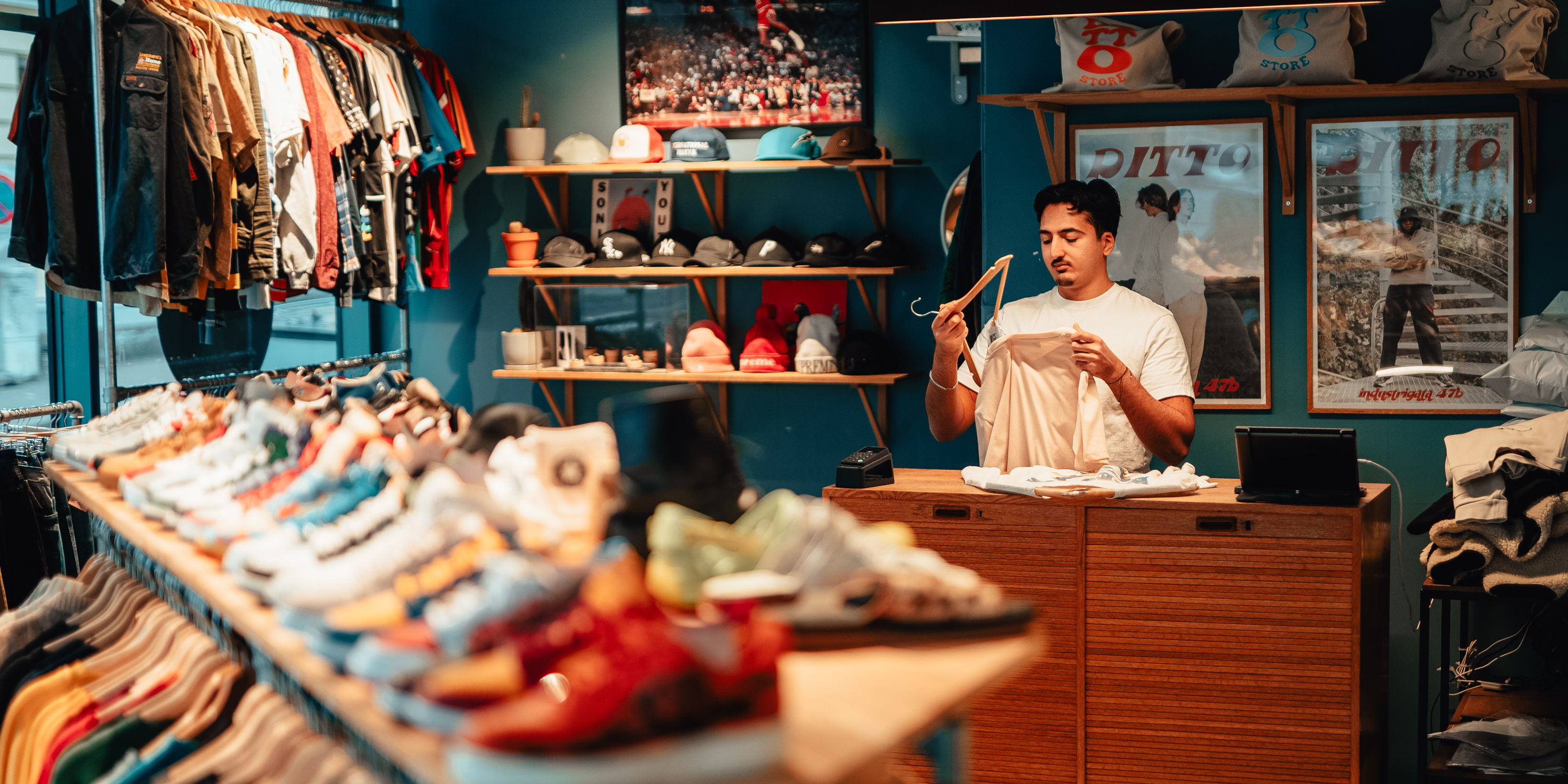 Klær og sko på hyller i en butikk, med en mann som står i kassen og bretter klær