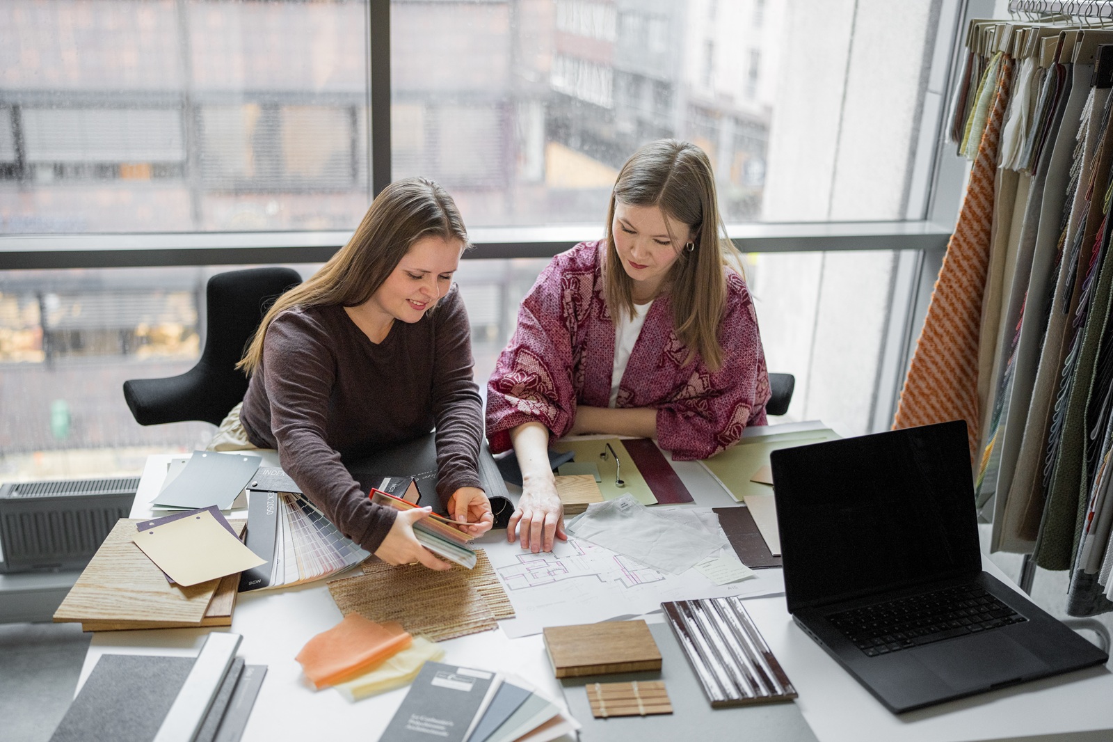 To studenter samarbeider rundt et bord med materialprøver, plantegninger og fargekart på studiet Interiørdesign og romarkitektur ved Fagskolen Kristiania.