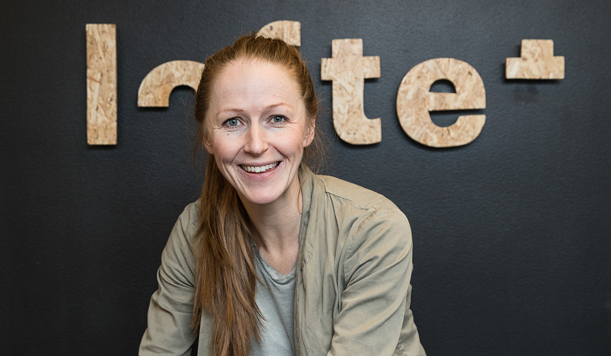 Faglig studieleder, Kristin Undheim, står foran loftet sin logo i lyst treverk. Veggen bak er sort. Kristin har på seg en grå cardiganjakke og har håret oppsatt i en hestehale. 