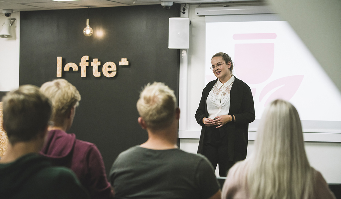 En student står foran en folkemengde på Loftet og forklarer et tema. Hun holder begge hendene samlet, og har de liggende i hverandre. Hun har på seg en hvit bluse og sort cardigan. 