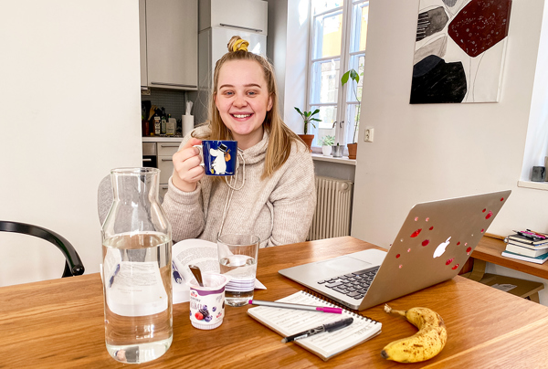 Foto av kvinnelig student ved kjøkkenbordet, som drikker kaffe mens hun jobber med en PC. På bordet ligger skriveblokk og banan.
