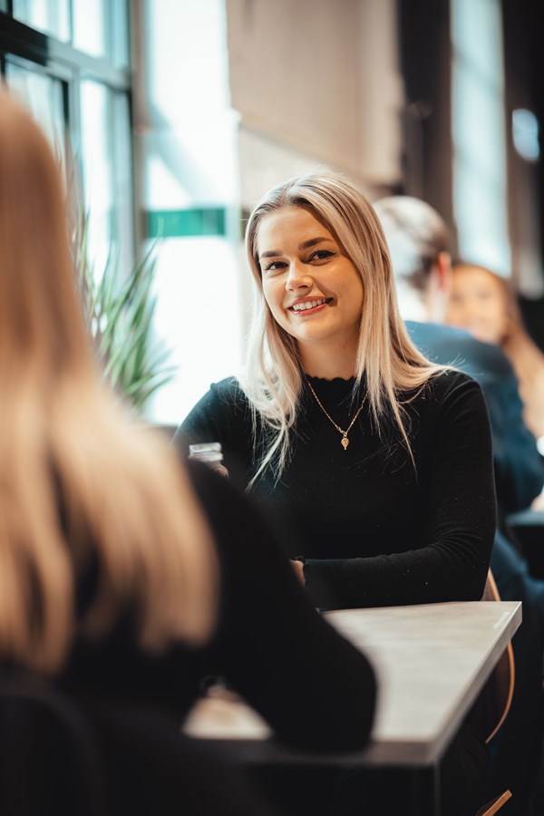 En person med lyst hår og en svart topp smiler til kamera, sitter ved et bord i et rom fylt med naturlig lys og andre personer i bakgrunnen.