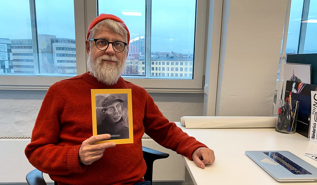 Mann med briller og skjegg holder opp en liten bok. Han sitter ved en pult.