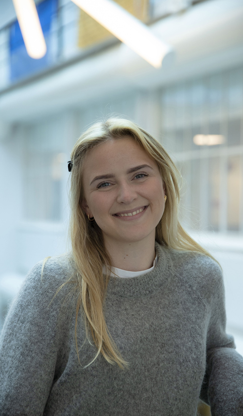 Kristiane Lunde studerer Music Business ved Fagskolen Kristiania. Hun lener seg på gelenderet i trappen på Campus Fjerdingen med foajeen i bakgrunnen. Kristiane har langt, blondt hår, mørke øyenbryn og blå øyne. Hun har på seg en sort hårspenne, gulløredobber og en hvit topp med en grå ullgenser over. 