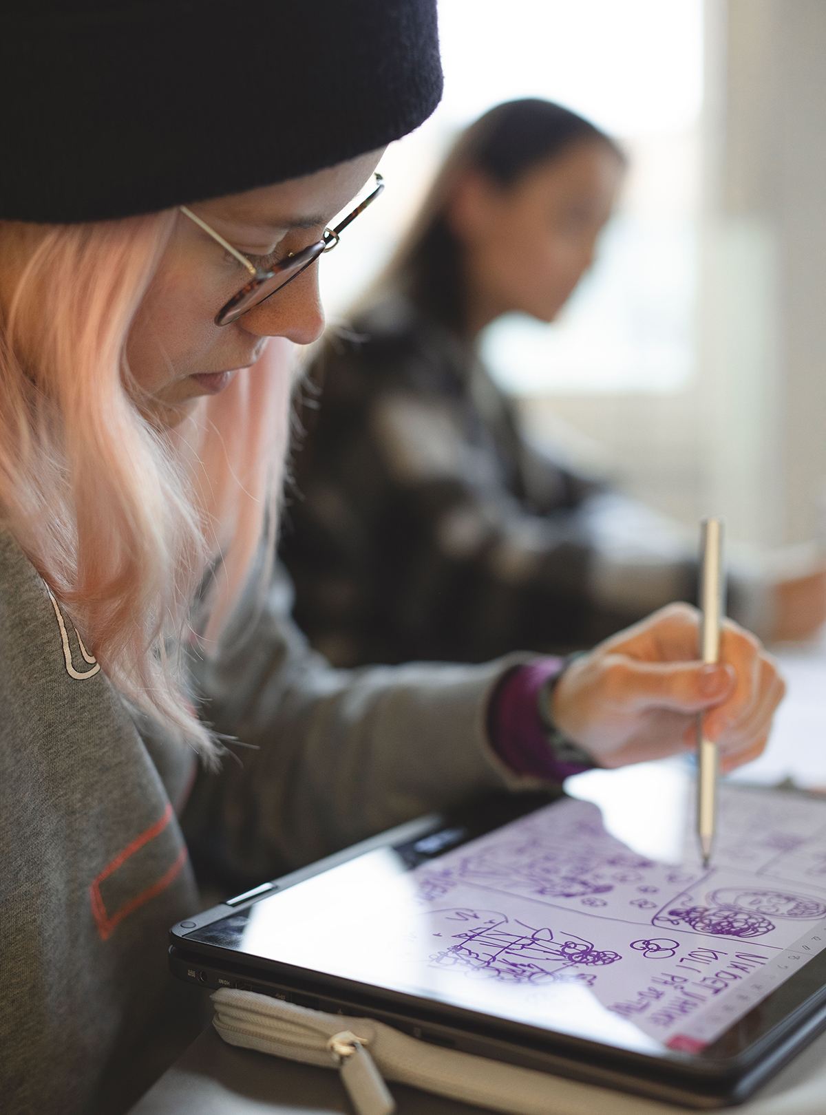 En student med lyserosa hår sitter og tegner på et digitalt tegnebrett. Hun ser konsentrert ut.