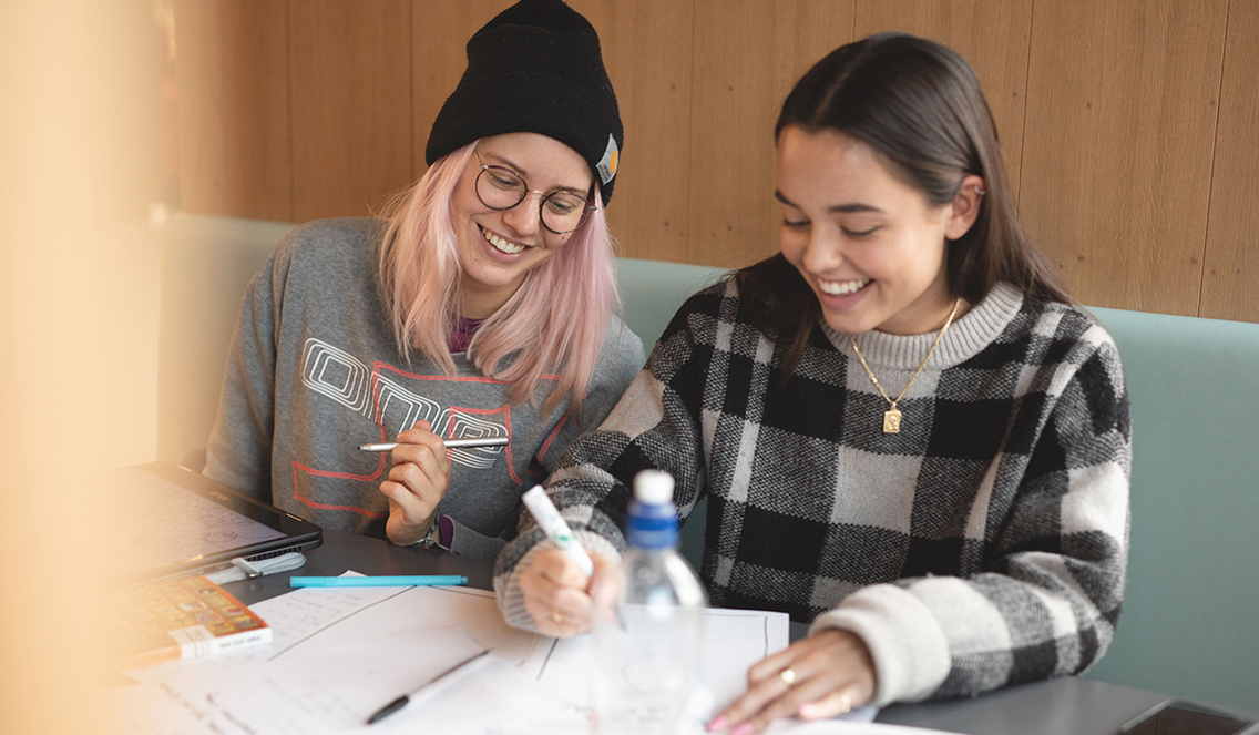 To studenter som studerer Art Direction sitter inne på et lite grupperom med trevegger og skisserer på ark som ligger på bordet foran dem. Hun ene har lyserosa hår. Hun har på seg en sort lue og har runde briller. Hun smiler og ser ned mot skissene på bordet. Den andre studenten har langt brunt hår og har et gullsmykke rundt halsen. Hun holder i en sort markeringstusj og skriver på skissen mens hun smiler og ser ned på tegningen. 