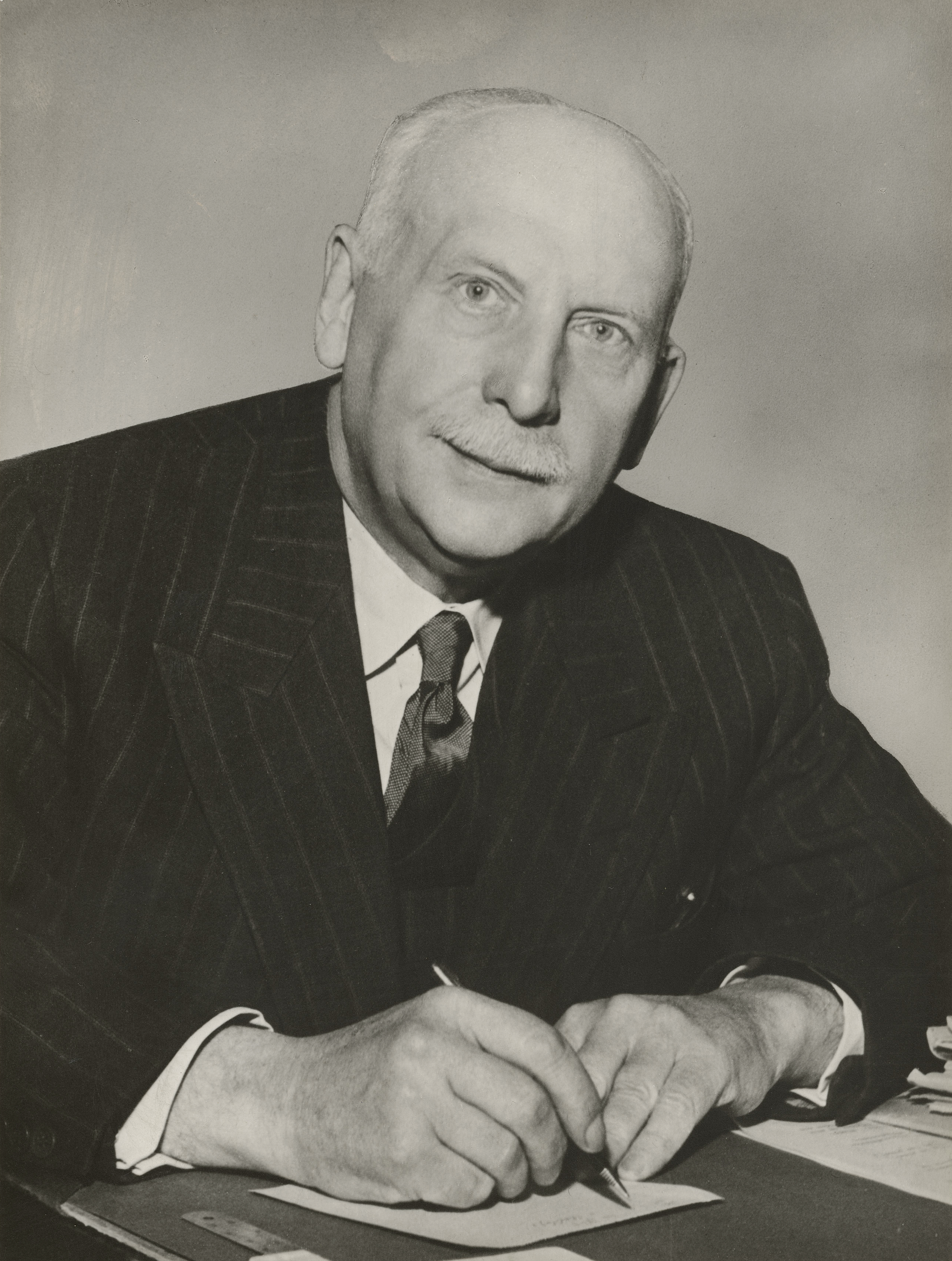 Bildet er av en eldre mann i stripete dress og slips med en liten bart. 