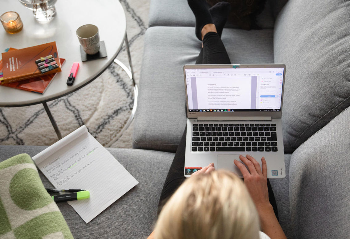 Illustrasjonsfoto tatt i fugleperspektiv av en nettstudent som sitter med utstrakte ben i en sofa med laptop på fanget og studerer. På sofaen og bordet ved siden av ligger det en notatblokk, skolebøker og markeringstusjer.