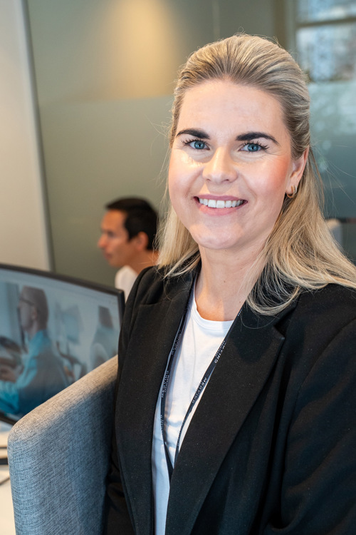 Portrett av Ragnhild som står på kontoret og smiler, bak henne er PC-skjerm og kollega. 