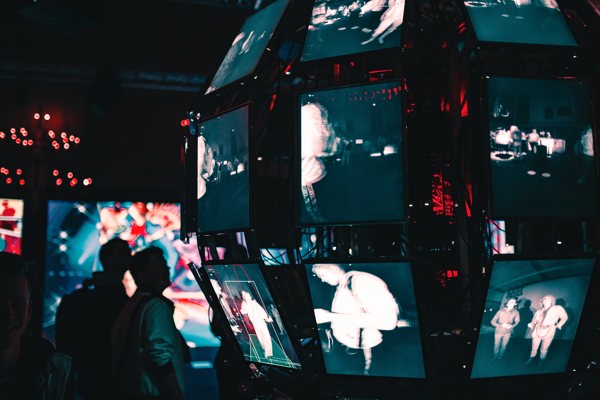 Two persons looking at multiple screens with humans on, in a dark room.