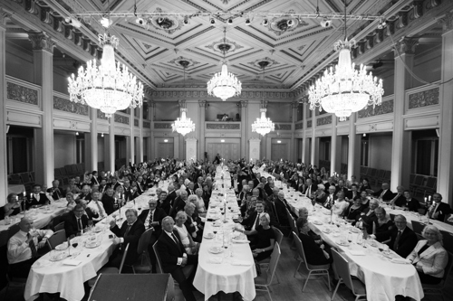 En flott festsal med store lysekroner er fyllt av fem langbord. Festpyntede mennesker sitter rundt alle bordene med hvite duker.