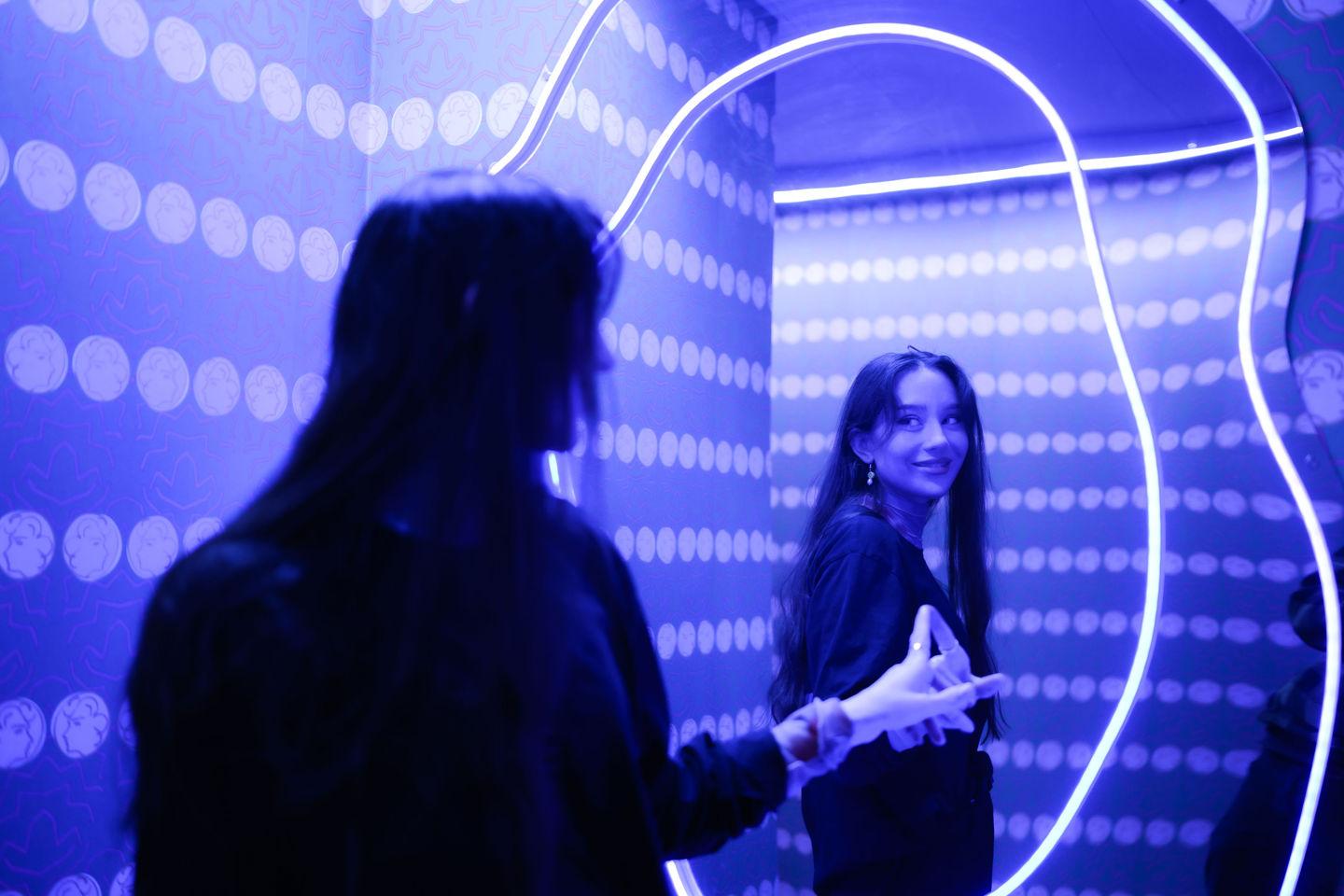 Bildet viser en kvinne foran et speil i et rom med blått lys. Speilbildet hennes er uskarpt i forgrunnen, mens hennes klare refleksjon smiler og poserer med hendene i speilet, som har en lysende neonring rundt seg. Bakgrunnen er dekorert med lyserende prikker, noe som bidrar til en livlig og moderne stemning.