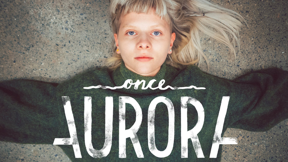 Aurora Aksnes ligger på bakken og ser inn i kamera. Hun har blondt hår med pannelugg og en grønn genser.