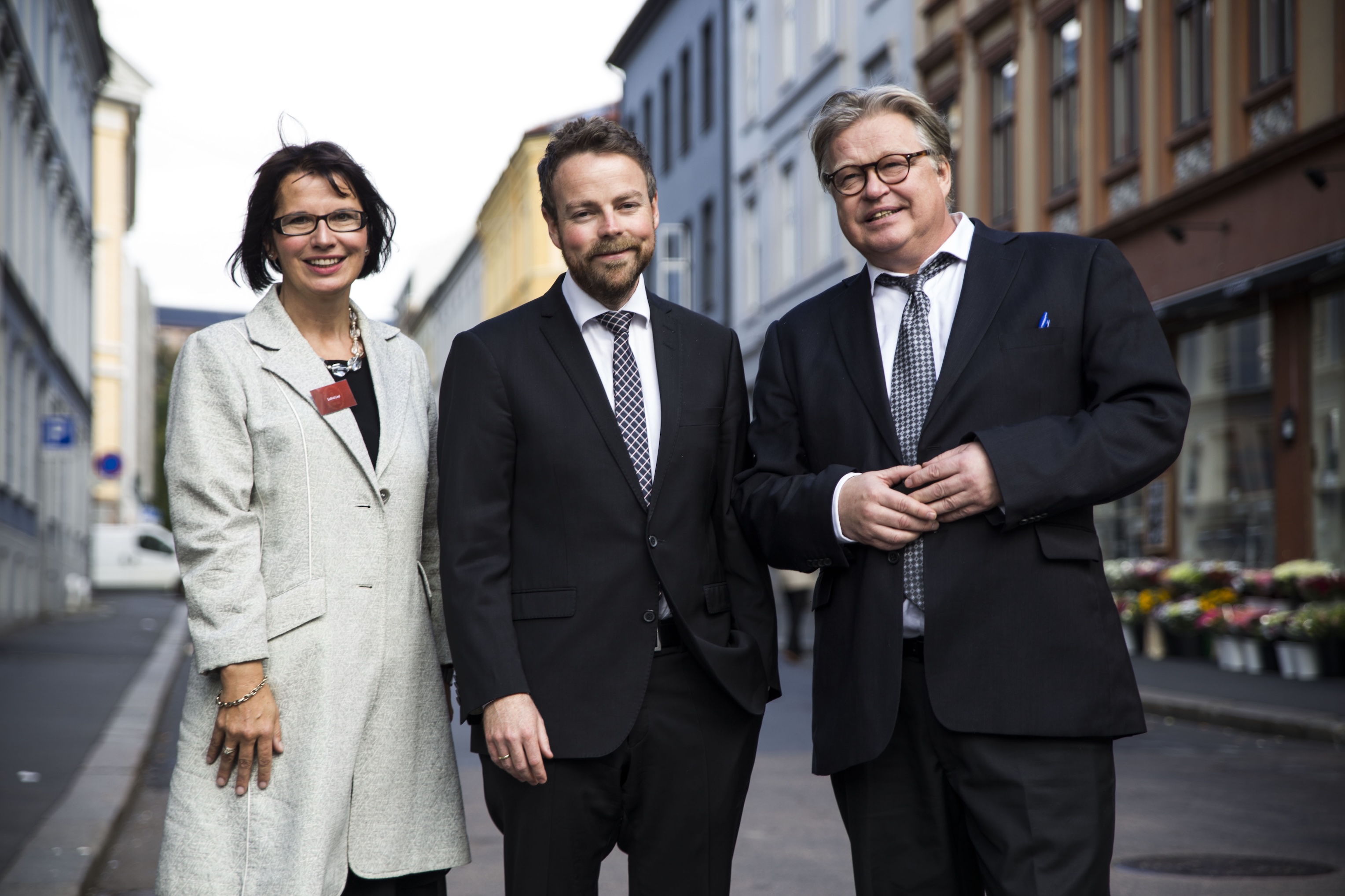 To menn i dress og en dame står i gaten og smiler til kameraet.