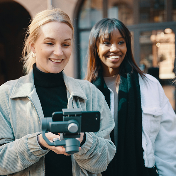 Foto av to kvinnelige studenter som står ved siden av hverandre. Den ene filmer med en mobil som er festet i en stabilisator.   