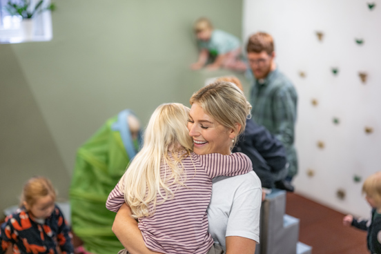 En voksen holder et barn i armene i et rom der flere barn leker rundt på ulike aktiviteter.