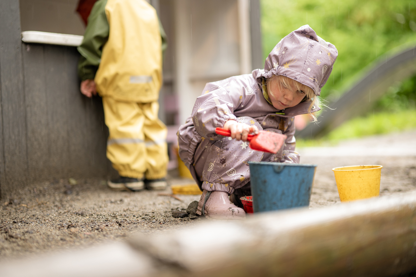 Barn som leker ute i sandområdet med bøtter og spade ved et lite lekehus