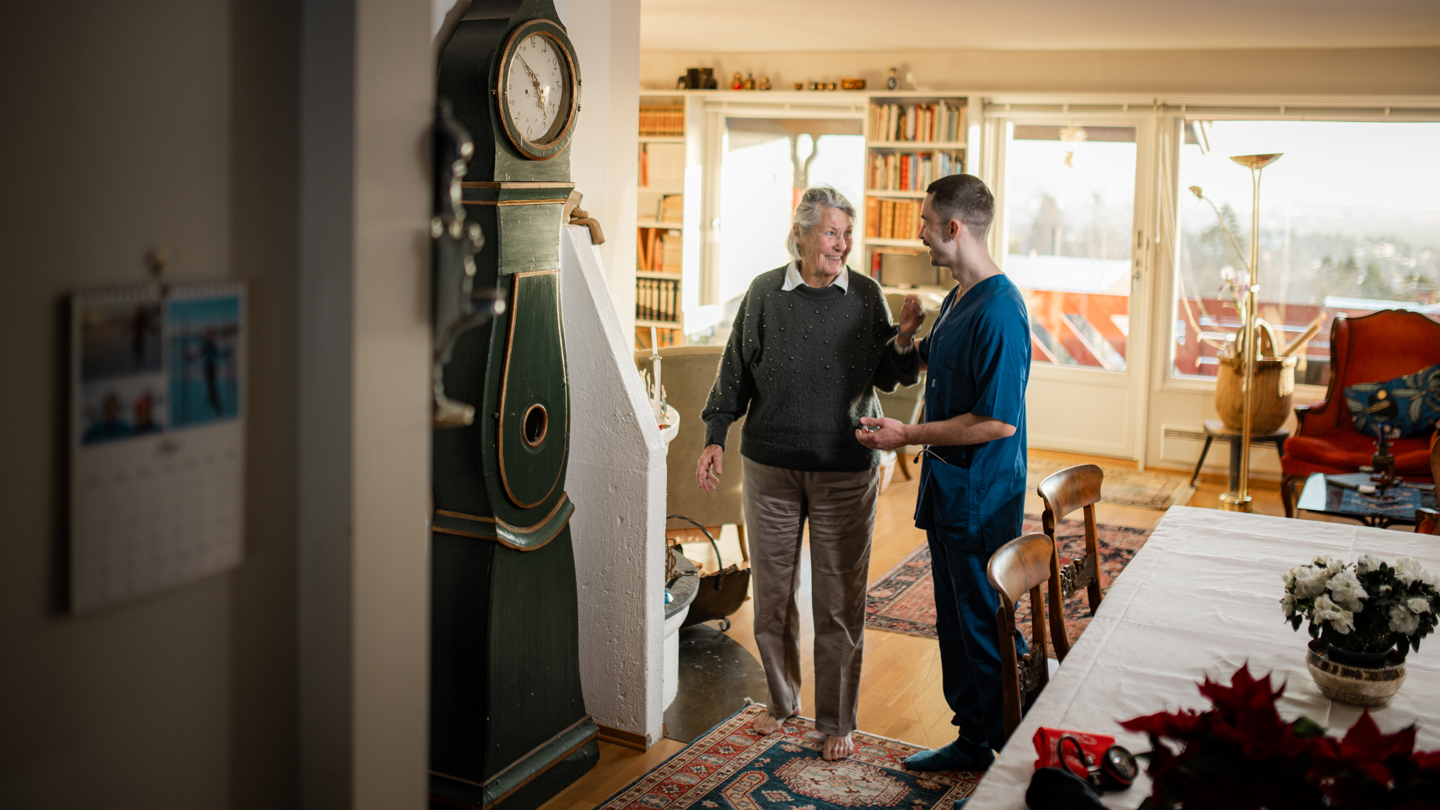 En helsearbeider står og snakker med en eldre person i en lys og hyggelig stue