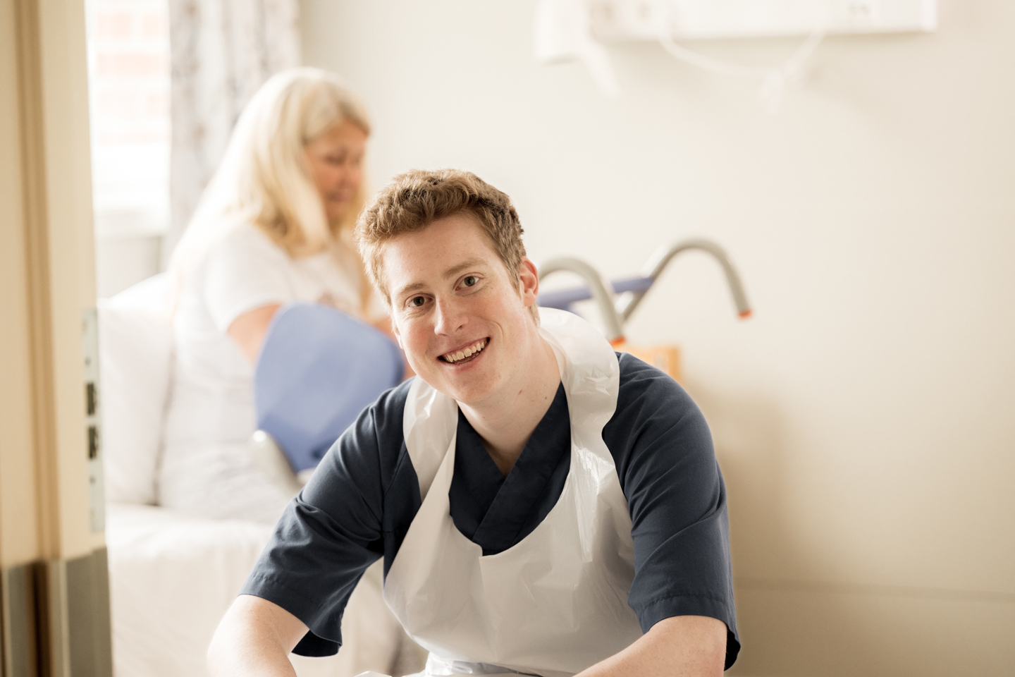 Helsefagarbeider smiler i kamera med pasient i bakgrunnen.