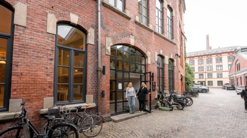 To personer står utenfor inngangen til en murbygning i en brosteinsbelagt gate med sykler parkert langs veggen.