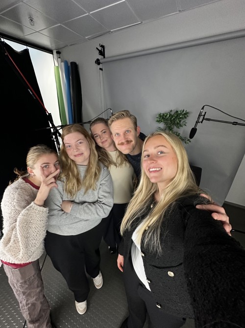 Gruppe på fem personer tar en selfie i et studio med fotoutstyr og mikrofon i bakgrunnen