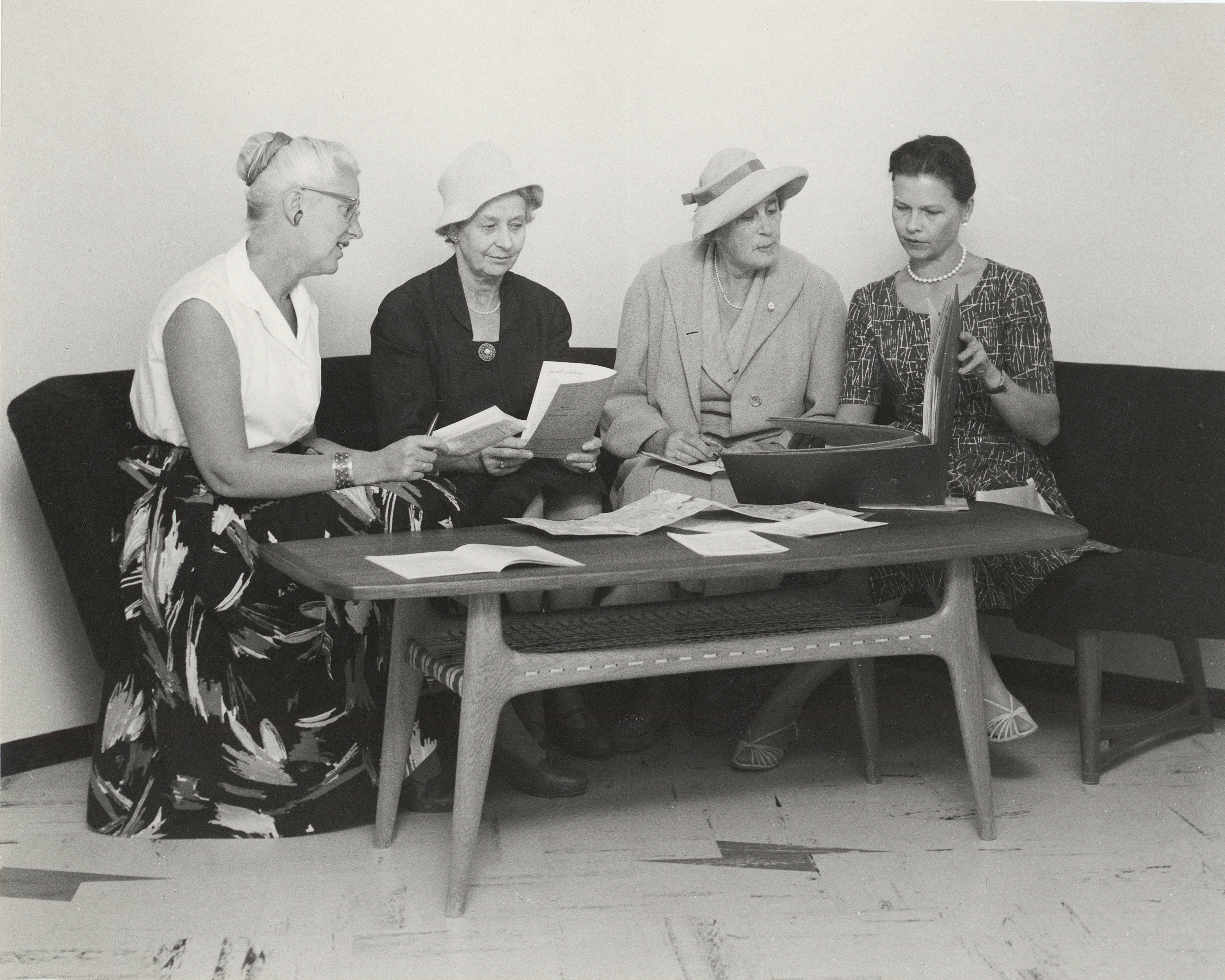 Fire pent antrukne damer fra 50-tallet sitter rundt et bord og leser ulike papirer. 