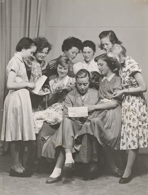 Mange unge damer og piker fra 1950-tallet med kjoler og pene frisyrer er samlet rundt noen papirer.