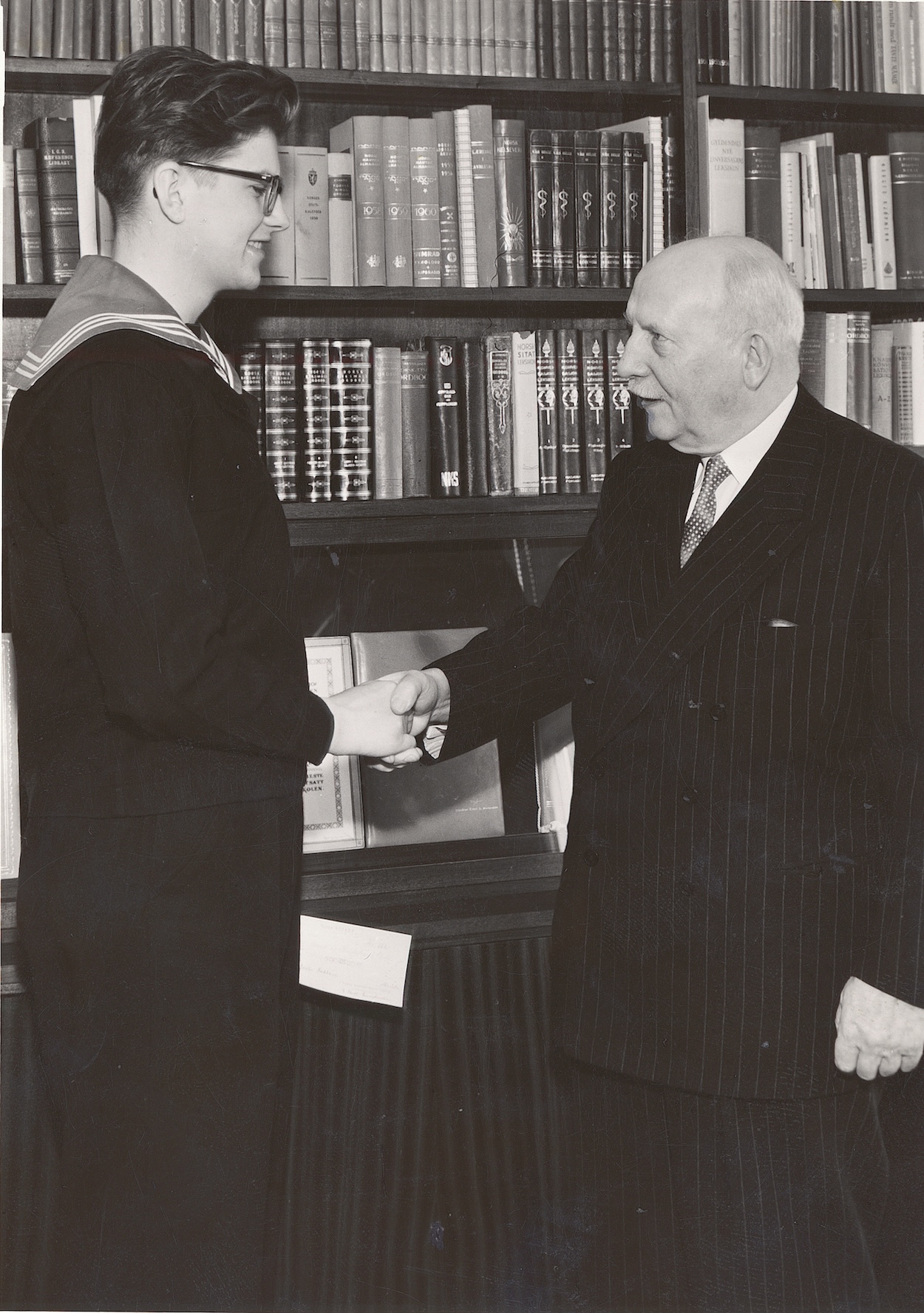 Bildet viser en ung mann i matrosdress og en eldre mann som tar hverandre i hånden.