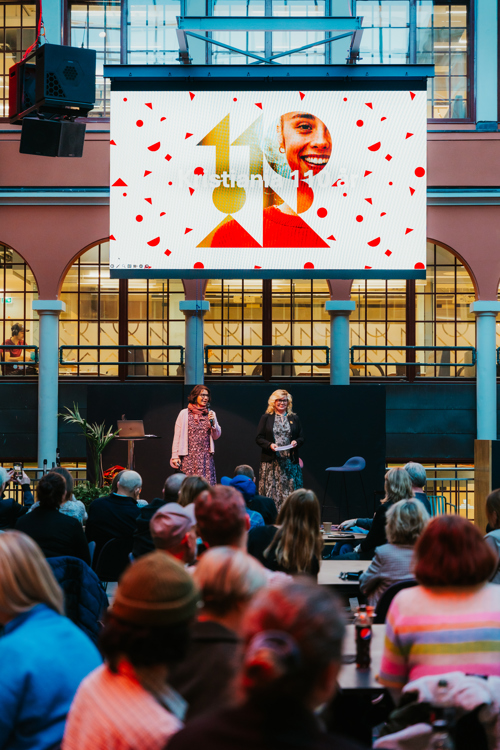 Bildet viser ryggen til mange mennesker som står og ser på to pent kledde damer som står oppe på en scene. Over dem henger et stort banner hvor det står: 110 år.