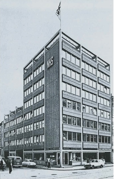 Foto av et kontorbygg fra 60-tallet med et par biler i gatene og fotgjengere på fortauet.