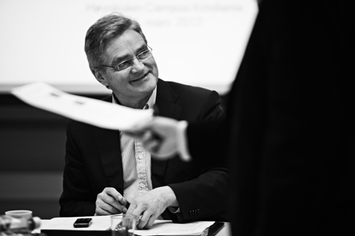 Bildet viser en smilende mann med briller. Han sitter ved et bord med noen papirer og en telefon foran seg. 