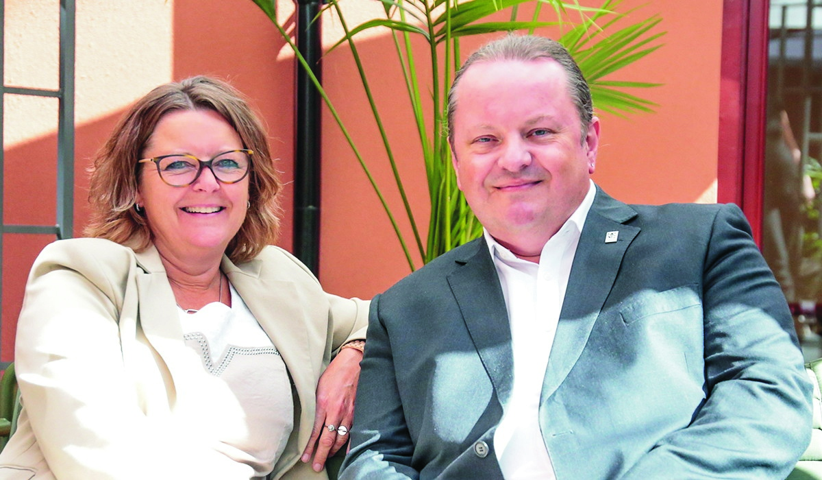 En kvinne med briller og en mann i dress sitter ved siden av hverandre og smiler.