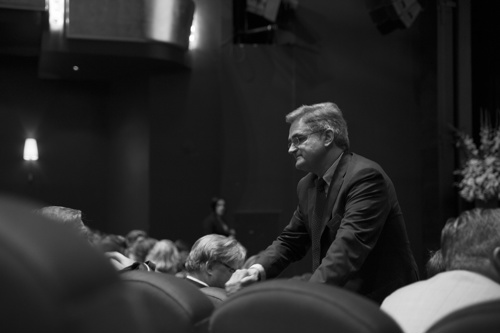 Bilde av en mann med briller og dress som reiser seg i en sal fullt av mennekser.