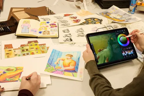 En person bruker tegnebrett. På bordet ligger det mange fargerike illustrasjoner