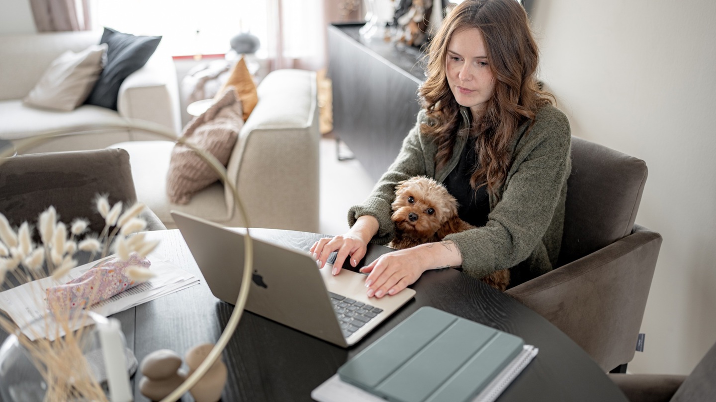 Student sitter ved et spisebord med en hund på fanget og en bærbar PC på bordet i et lyst hjemmemiljø.